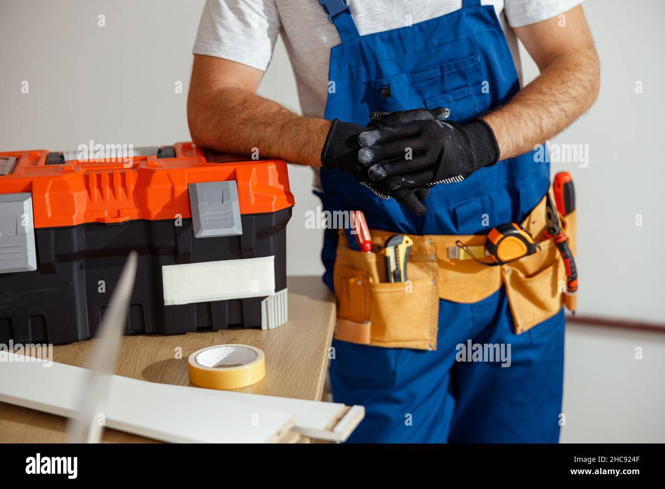 Scatto corto del tecnico in guanti uniformi e protettivi in posa per la telecamera in ambienti interni, appoggiati su una cassetta degli attrezzi Foto Stock