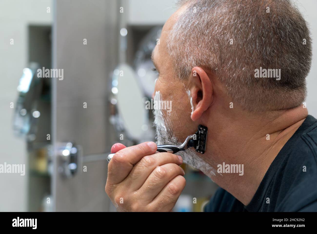 Uomo maturo dai capelli grigi che restyling la sua barba in casa utilizzando il rasoio Foto Stock