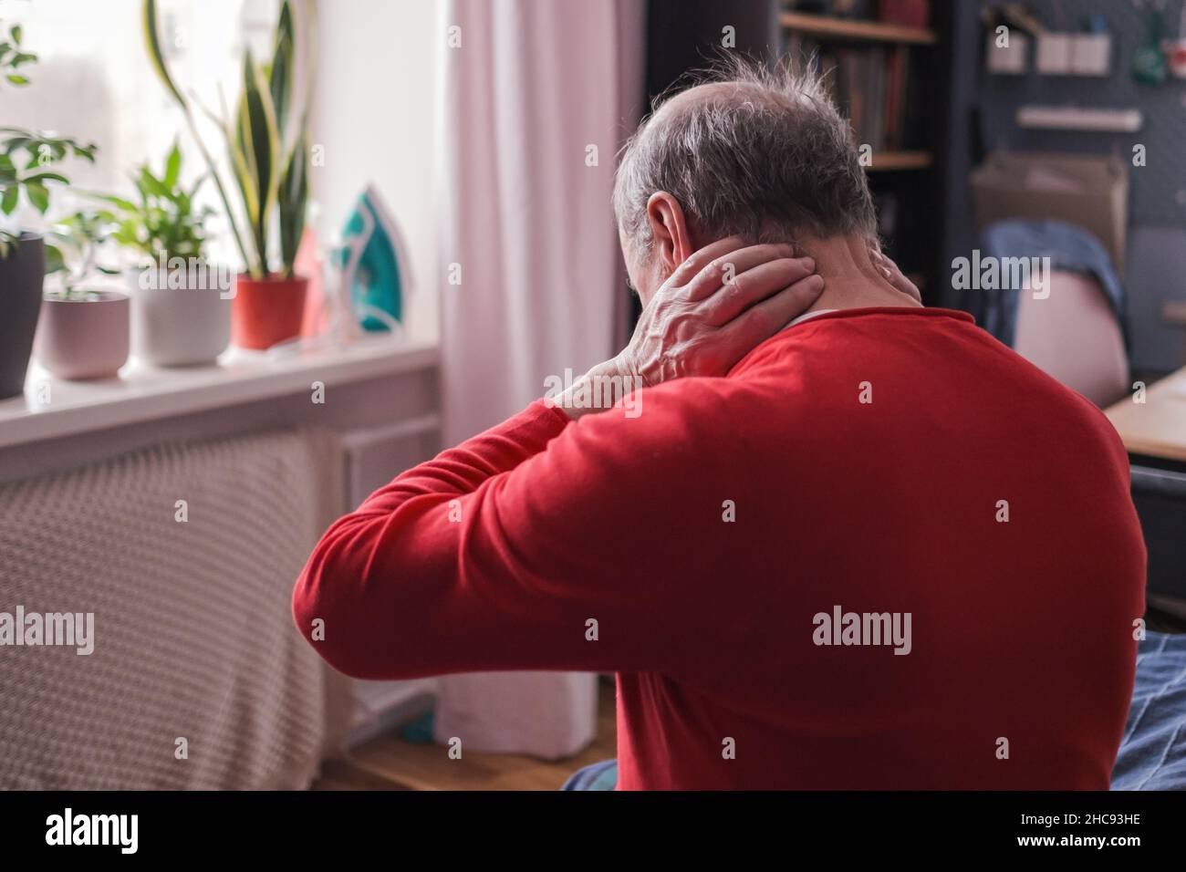 Uomo anziano frustrato in maglione rosso che tiene la mano sul collo seduto sul divano Foto Stock Uomo anziano frustrato in maglione rosso che tiene la mano sul collo seduto sul divano Foto Stock