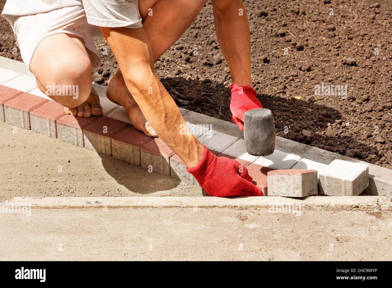 Un giovane lavoratore stende le lastre di pavimentazione in una luminosa giornata estiva di sole su una base di sabbia preparata sul marciapiede. Spazio di copia. Foto Stock