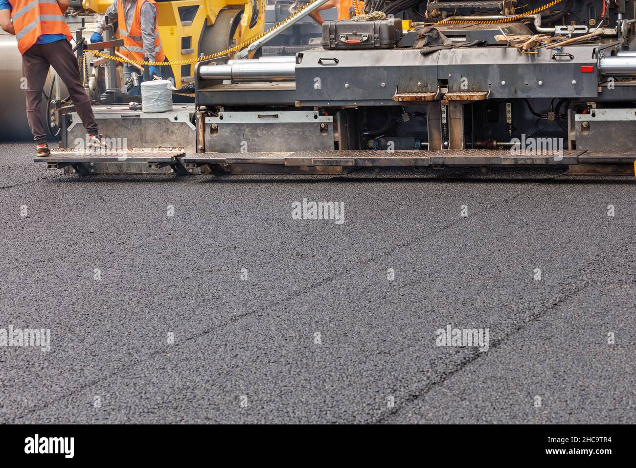 Un'asfaltatrice industriale stesa uno strato di nuovo asfalto caldo sulla carreggiata. Gli operatori controllano l'uniformità e l'altezza della pavimentazione. Spazio di copia. Foto Stock