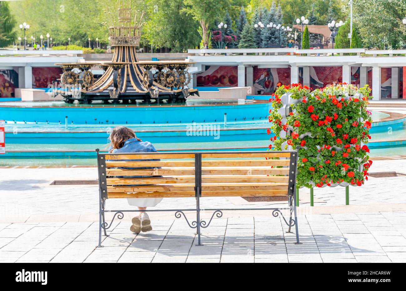 Vista posteriore di una donna seduta sulla panchina di fronte alla Fontana nel primo Parco del Presidente - un punto di riferimento urbano ad Almaty, Kazakhstan Foto Stock