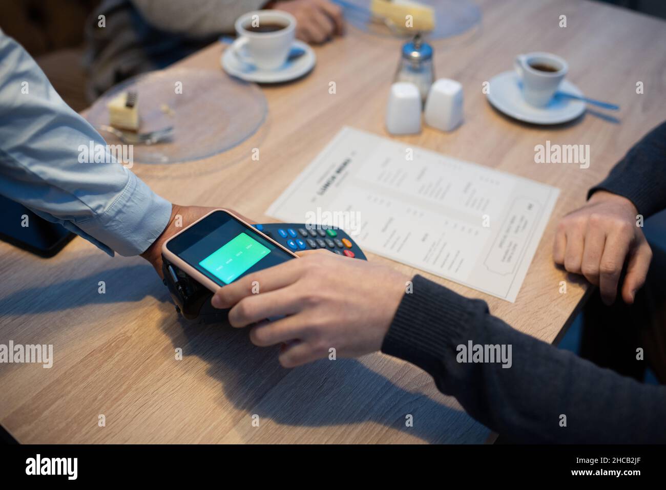 Giovane uomo che tiene smartphone sopra il terminale di pagamento tenuto dalla mano della cameriera mentre si siede da un tavolo di legno in un caffè Foto Stock