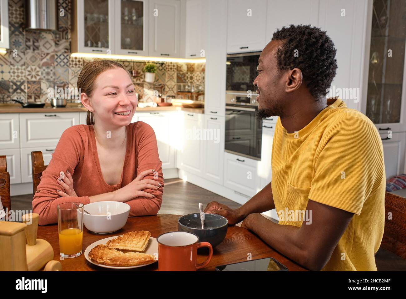 Felice giovane coppia interculturale chiacchierando con la prima colazione al mattino mentre si siede da tavolo da cucina in legno Foto Stock