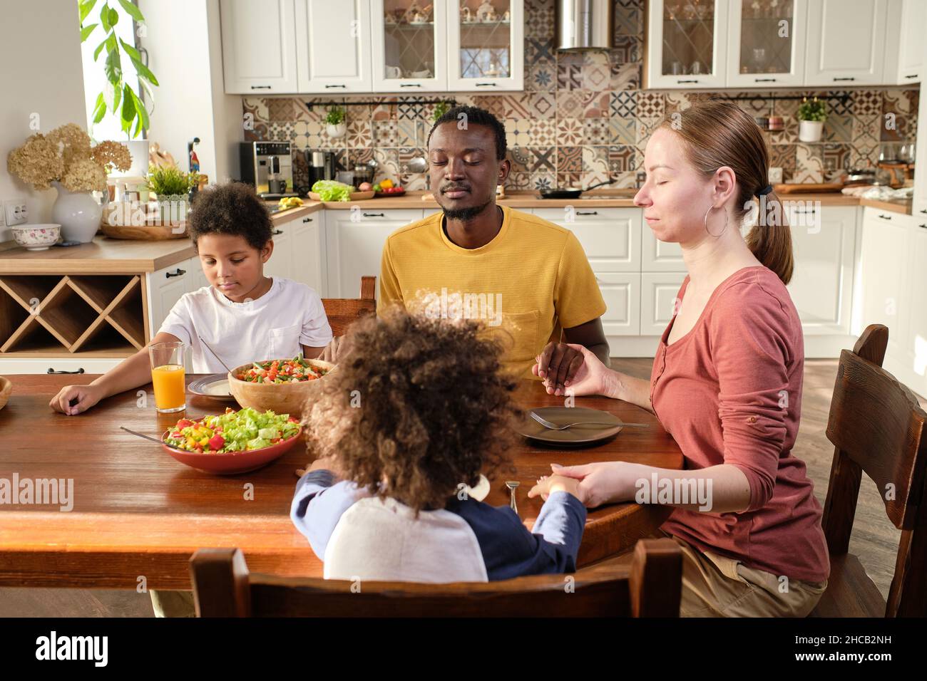 Giovane famiglia cristiana di quattro persone seduto al tavolo da cucina e tenuto con le mani mentre pregava prima di cena Foto Stock