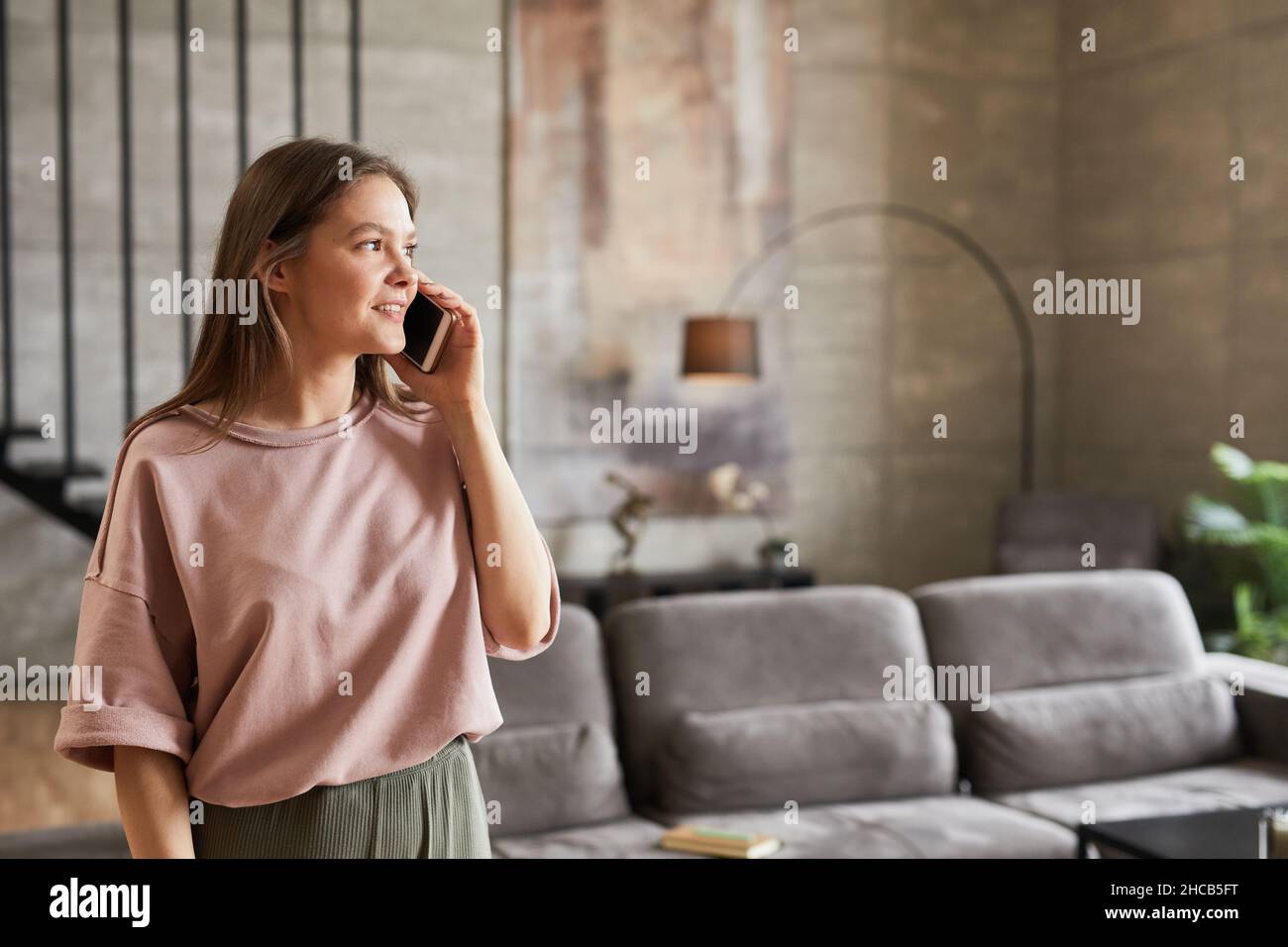 Giovane donna che ha una conversazione sul telefono cellulare e sorride mentre si trova nella stanza di casa Foto Stock