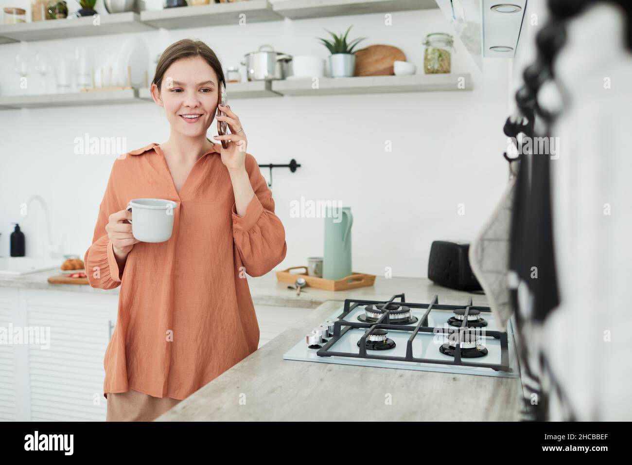 Giovane donna che ha una conversazione sul telefono cellulare mentre beve il caffè al mattino in cucina Foto Stock
