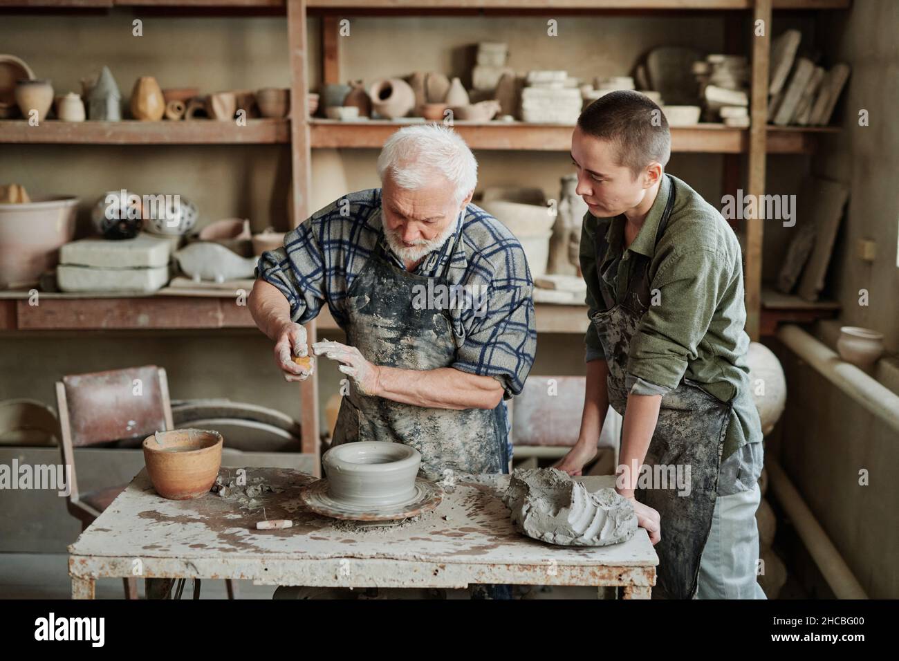 Uomo anziano che mostra il processo di fabbricazione dei vasi di ceramica al suo assistente nel laboratorio di ceramica Foto Stock