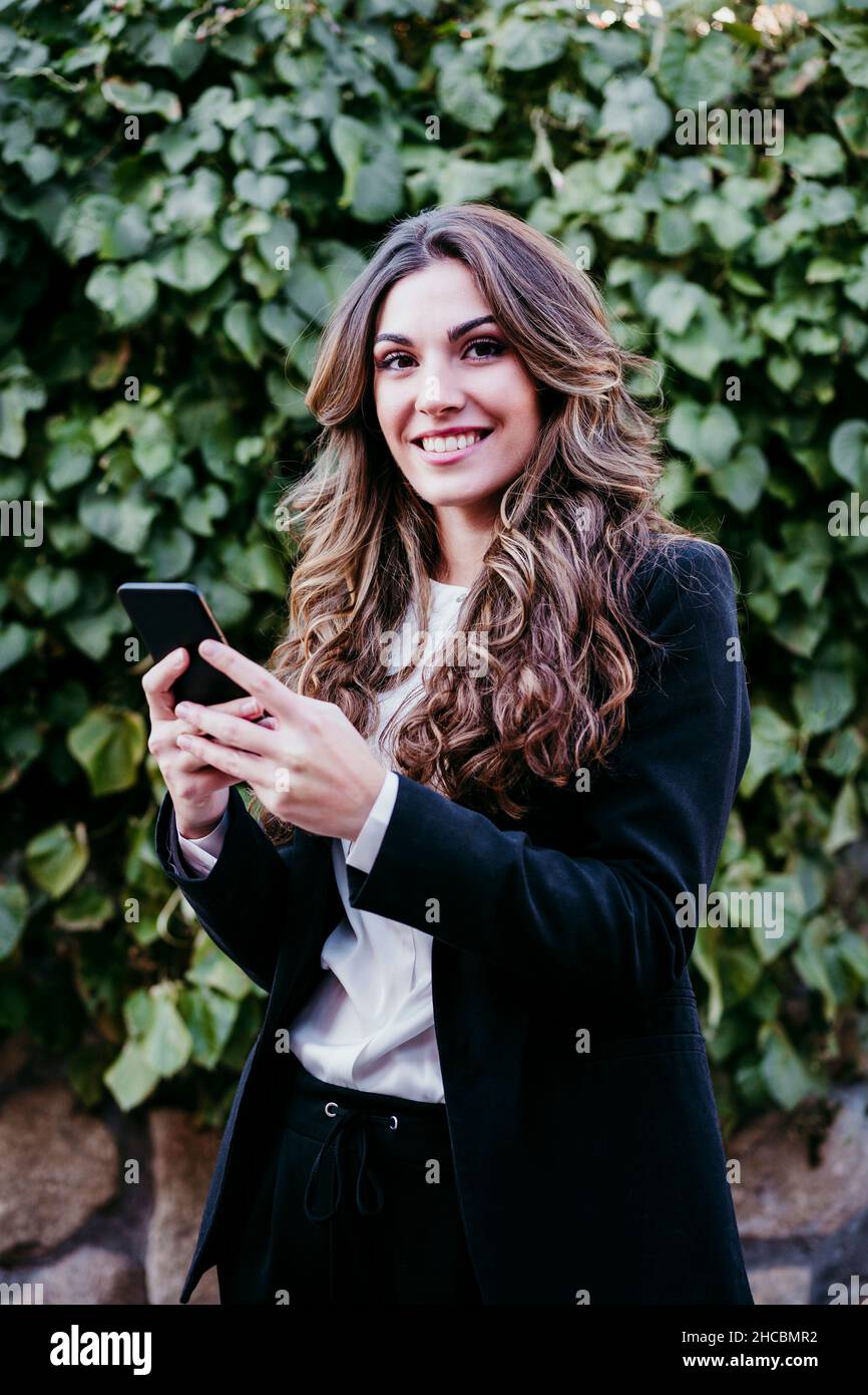Donna d'affari con capelli ondulati che tengono smartphone di fronte alla siepe Foto Stock