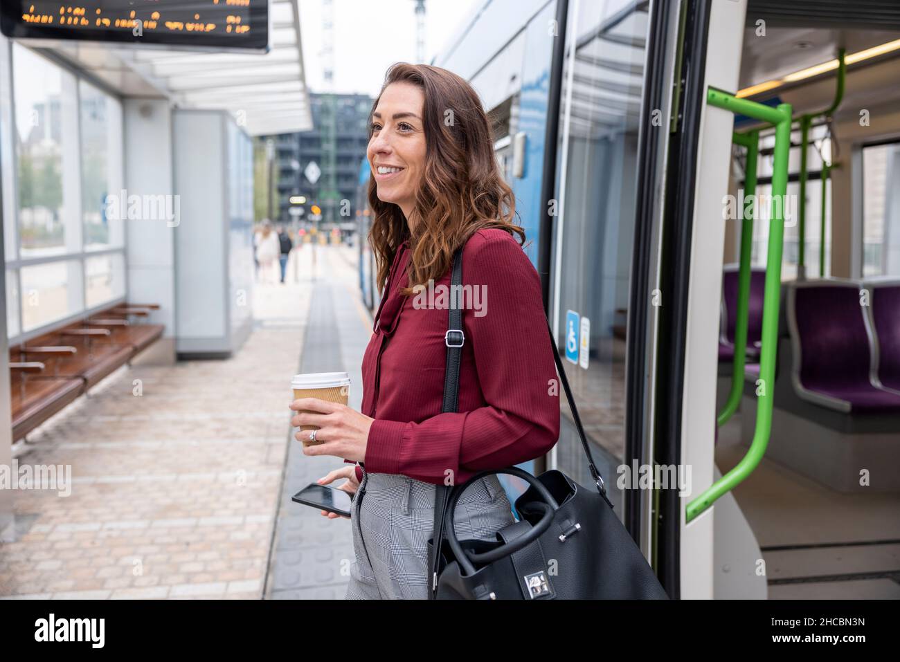 Donna d'affari sorridente che tiene la tazza usa e getta e il telefono cellulare sbarcare dal treno alla stazione Foto Stock