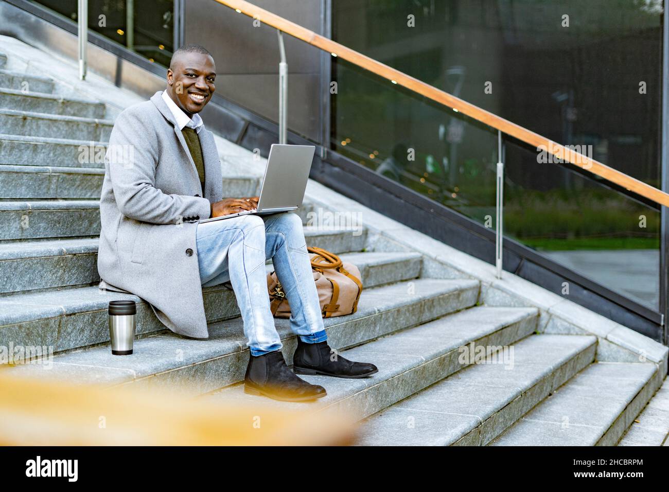 Uomo d'affari sorridente che tiene un contenitore di bevande isolato seduto con un computer portatile sui gradini Foto Stock