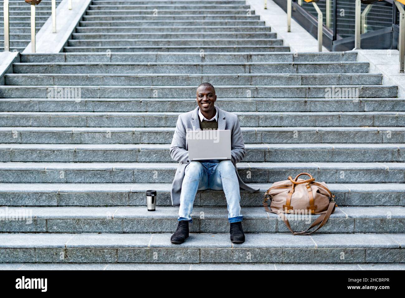 Uomo d'affari sorridente con borsone e contenitore per bevande isolato utilizzando un computer portatile seduto su gradini Foto Stock