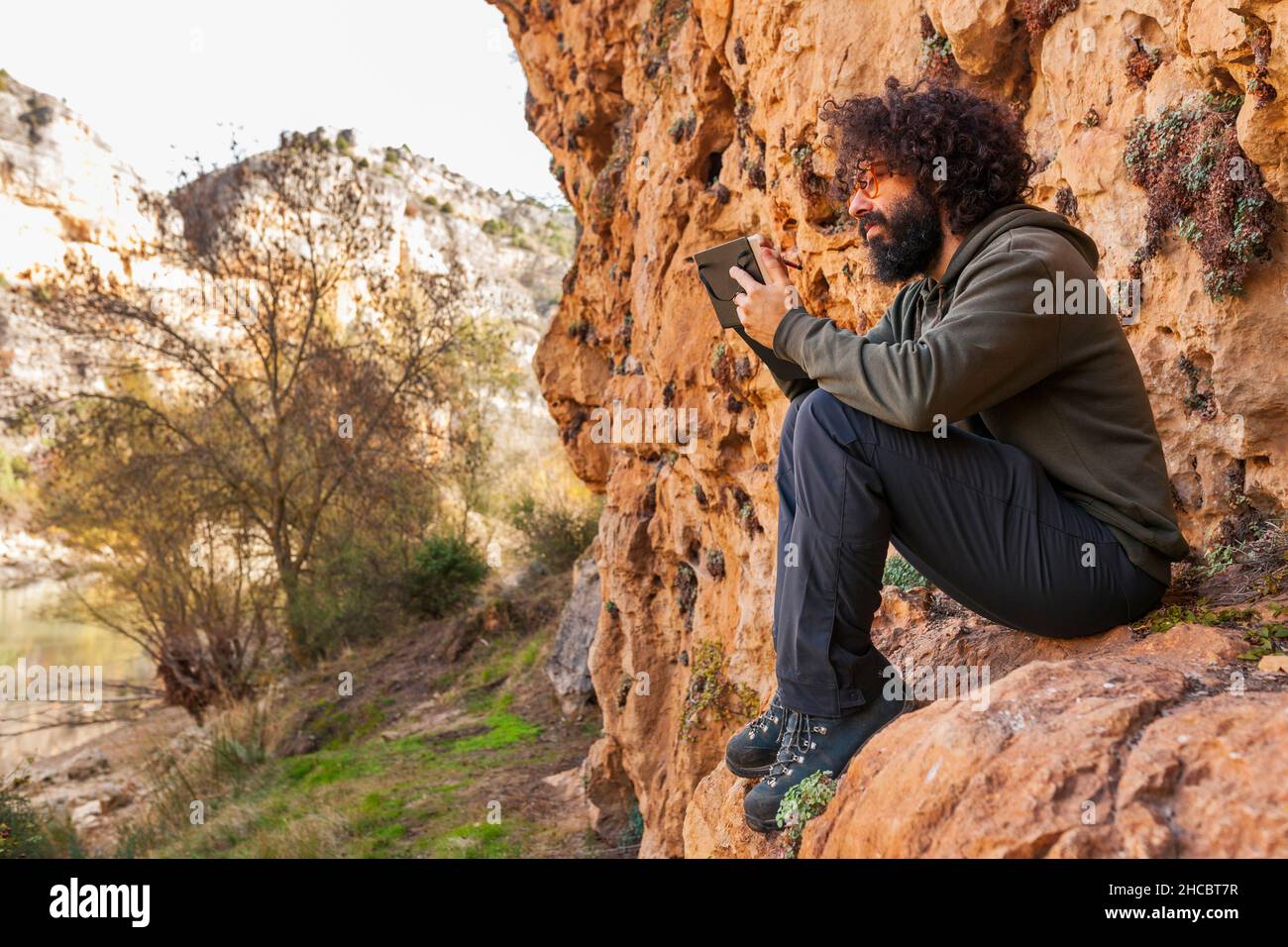 Uomo che si disegna in diario seduto su rocce vicino alla montagna Foto Stock