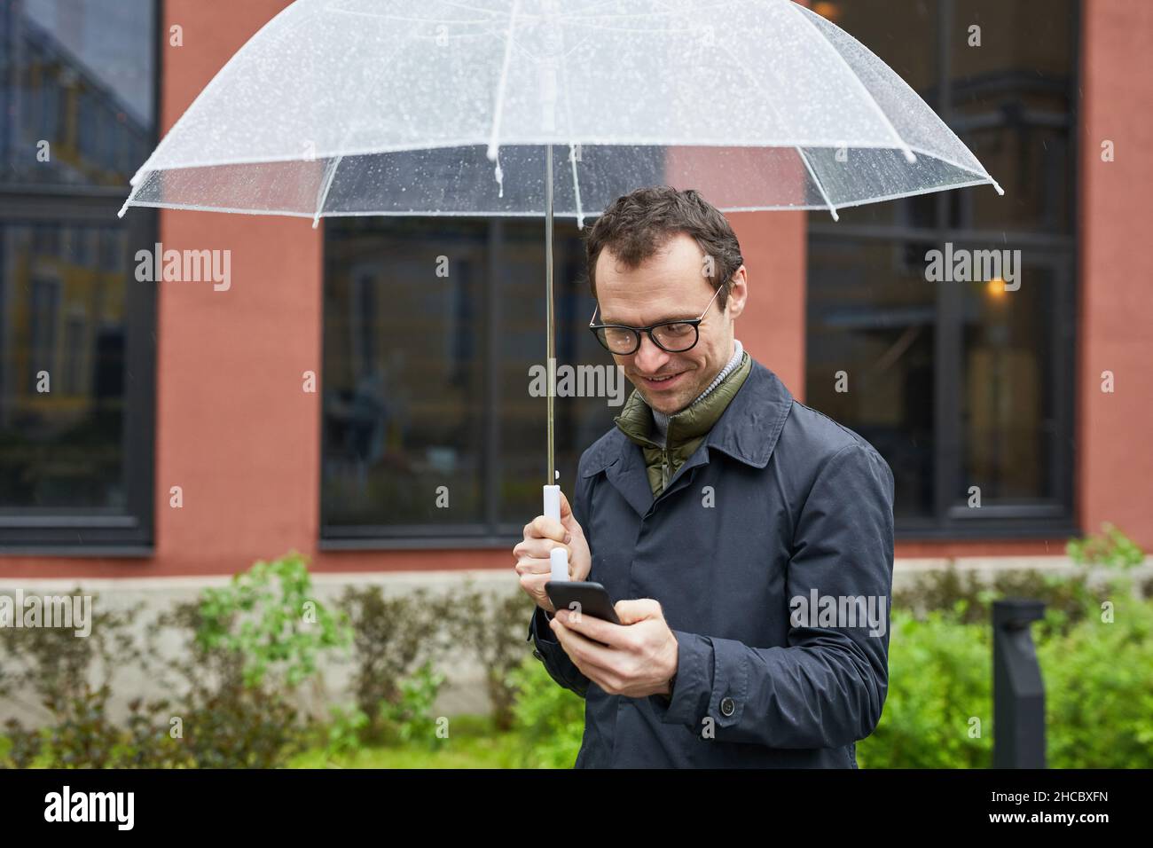 Scatto orizzontale medio di gioioso uomo caucasico indossando occhiali da vista trascorrere il tempo all'aperto in piedi sotto ombrello in pioggia surf Internet su smartphon Foto Stock