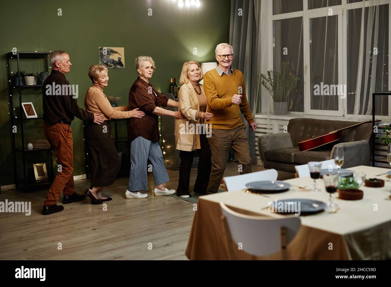 Due vecchi e tre donne che ballano e si divertono insieme in un soggiorno decorato con luci durante le riunioni di amici di festa Foto Stock