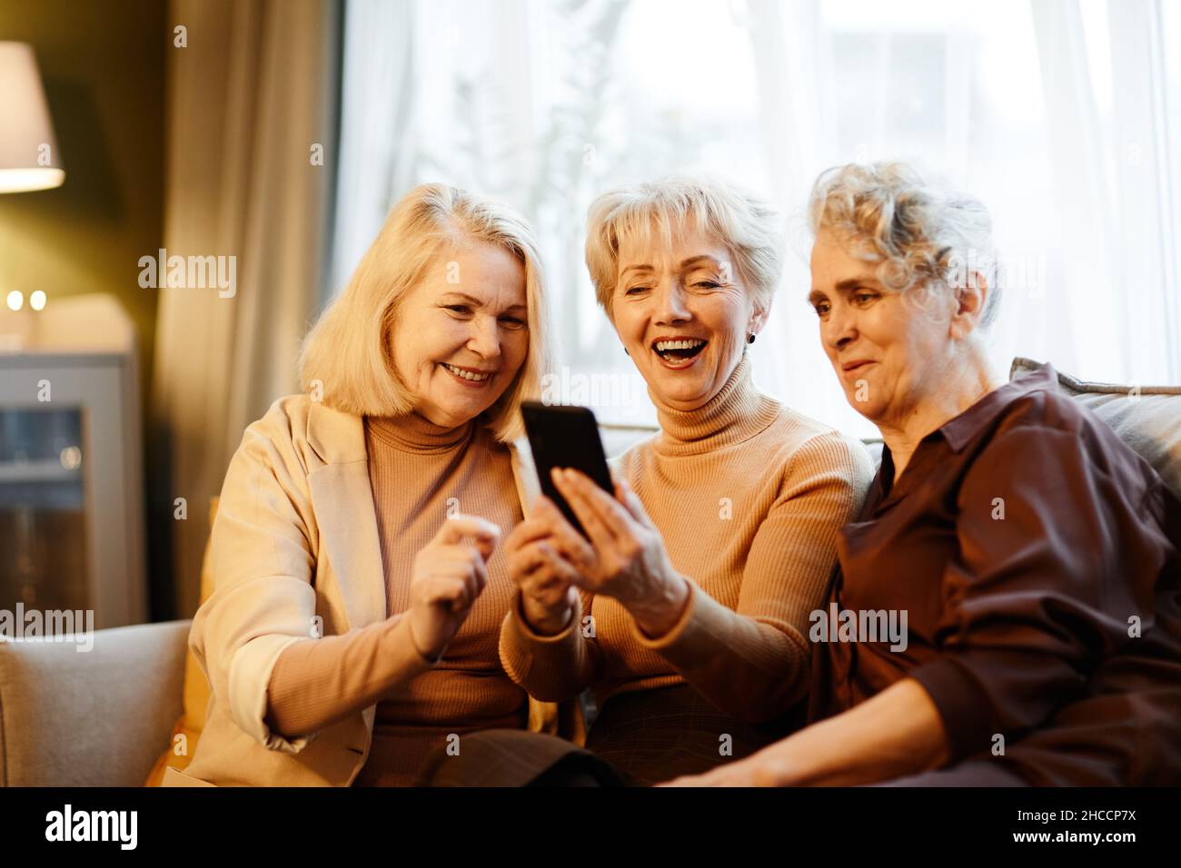 Gruppo di tre donne caucasiche di età felice seduti in soggiorno contro finestra e utilizzando smartphone Foto Stock