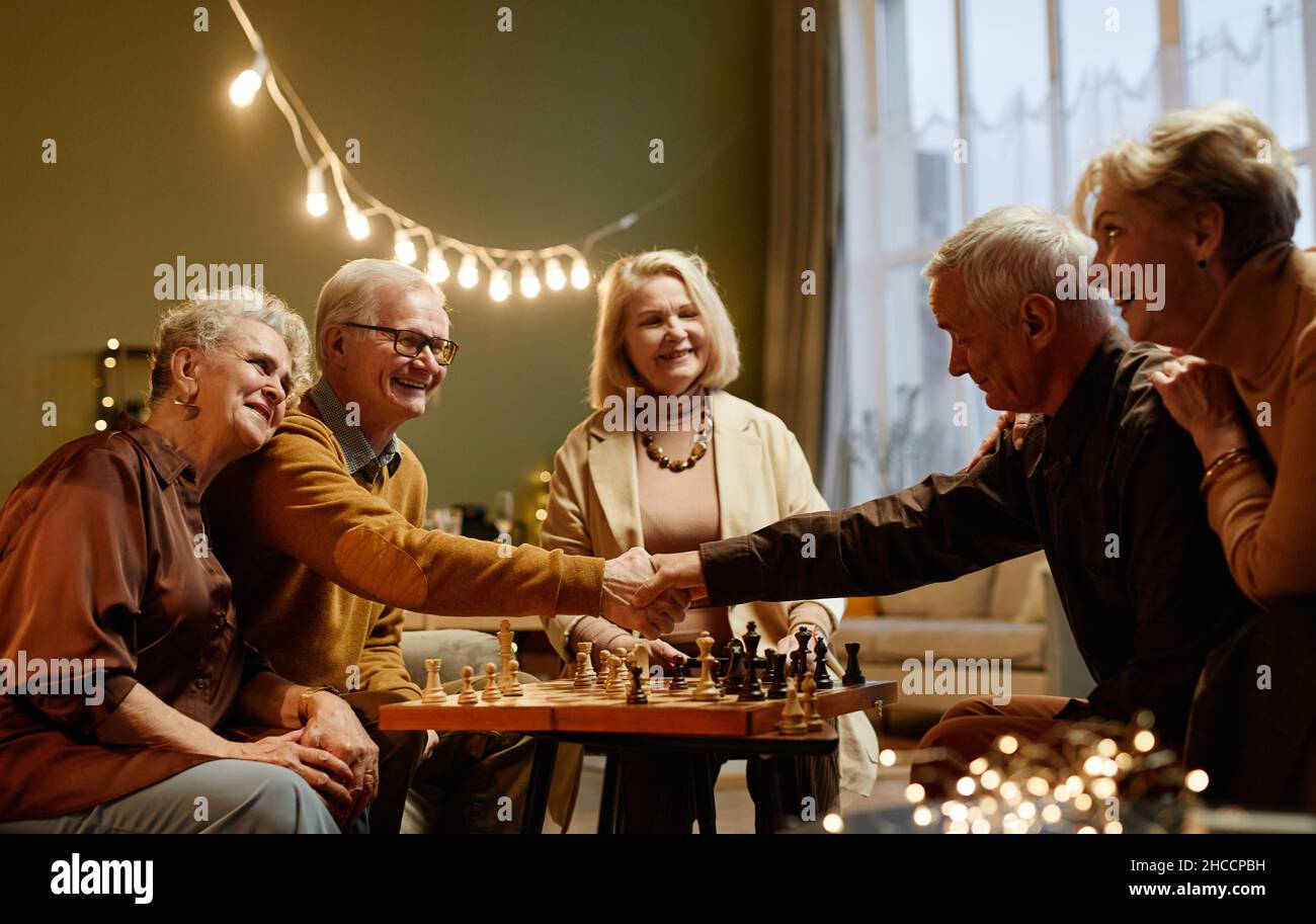 Due uomini caucasici di età che scuotono la mano dopo aver terminato il gioco degli scacchi, mentre tre donne anziane si siedono nelle vicinanze e guardano Foto Stock