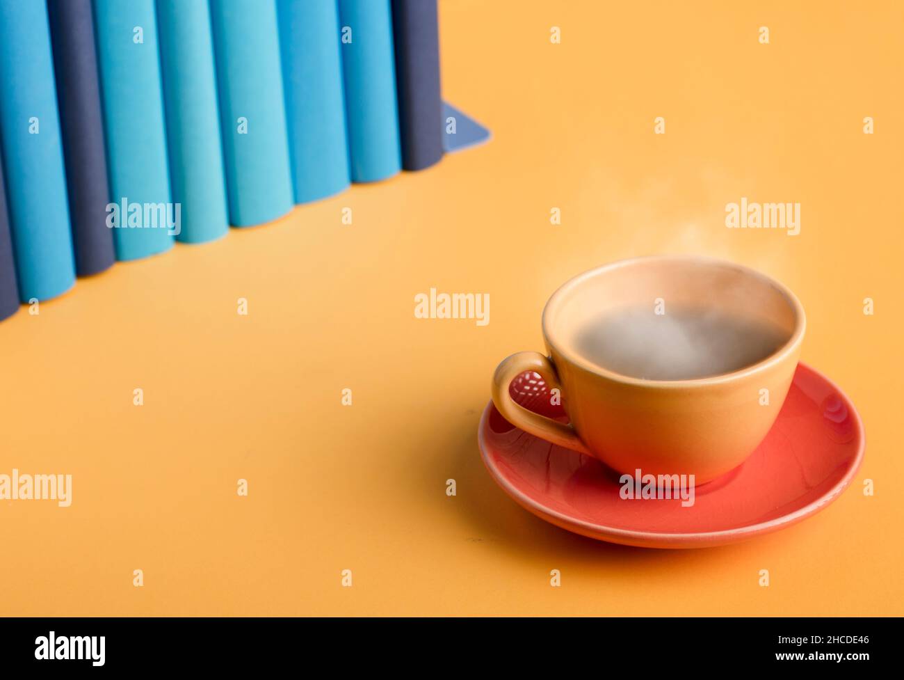 Tazza gialla di caffè o tè con vapore caldo, su un tavolo giallo, con libri blu e blu chiaro. Foto Stock