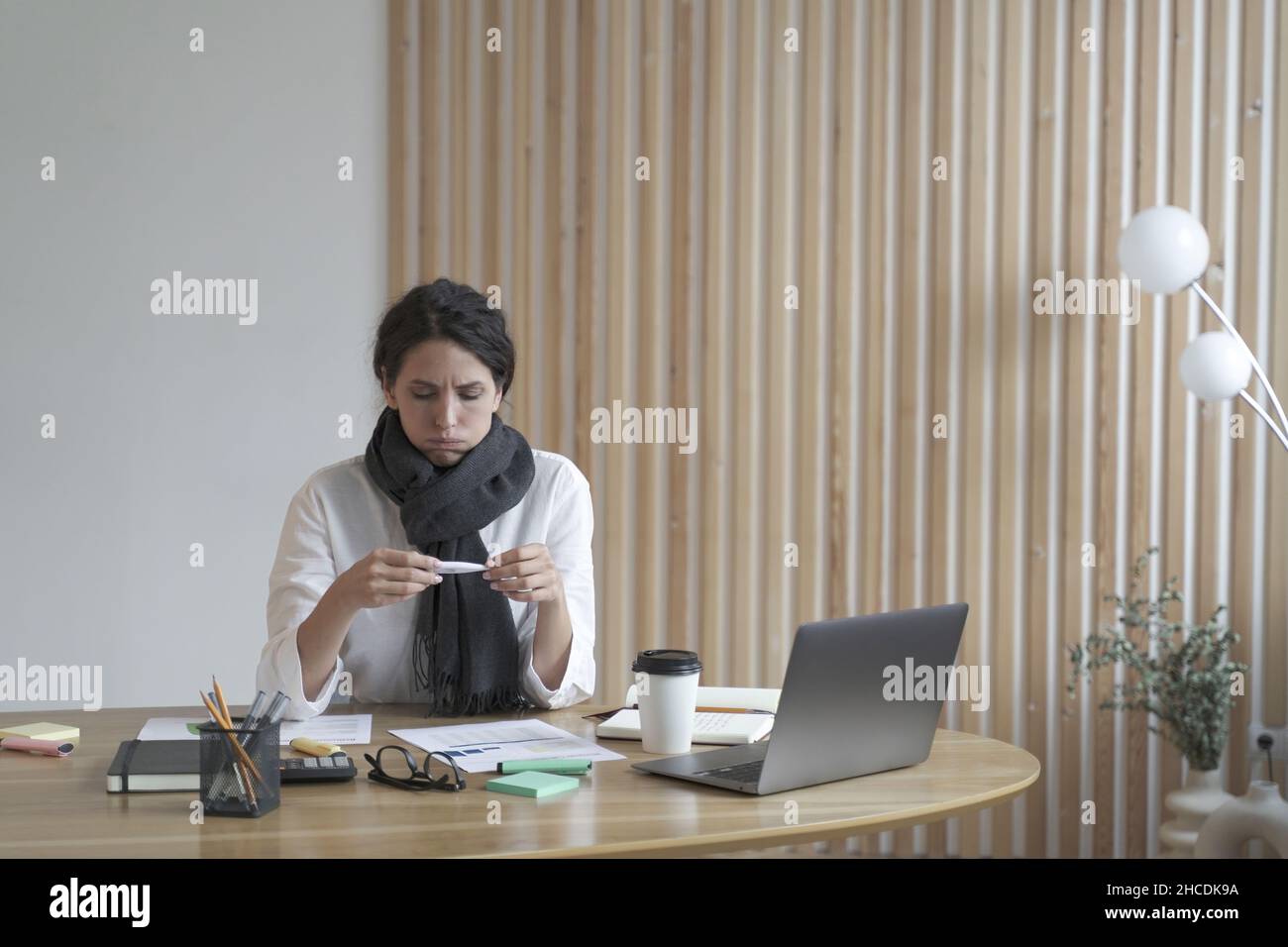 Malata giovane ispanica in sciarpa intorno al collo guardando il termometro elettronico sul posto di lavoro Foto Stock