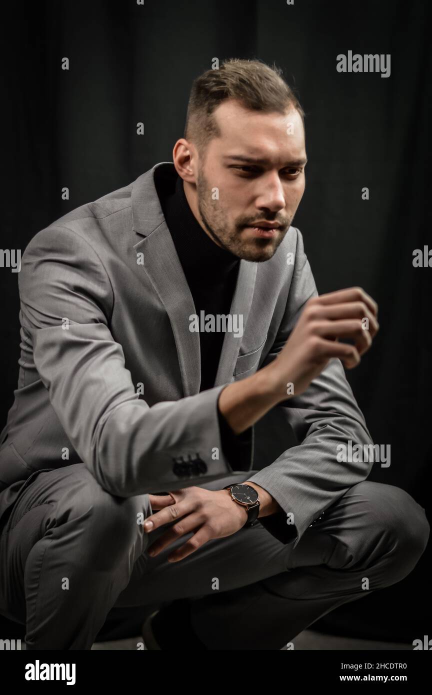 ritratto di un giovane uomo in un abito grigio e un dolcevita nero, accoccolato a terra con un aspetto intatto Foto Stock
