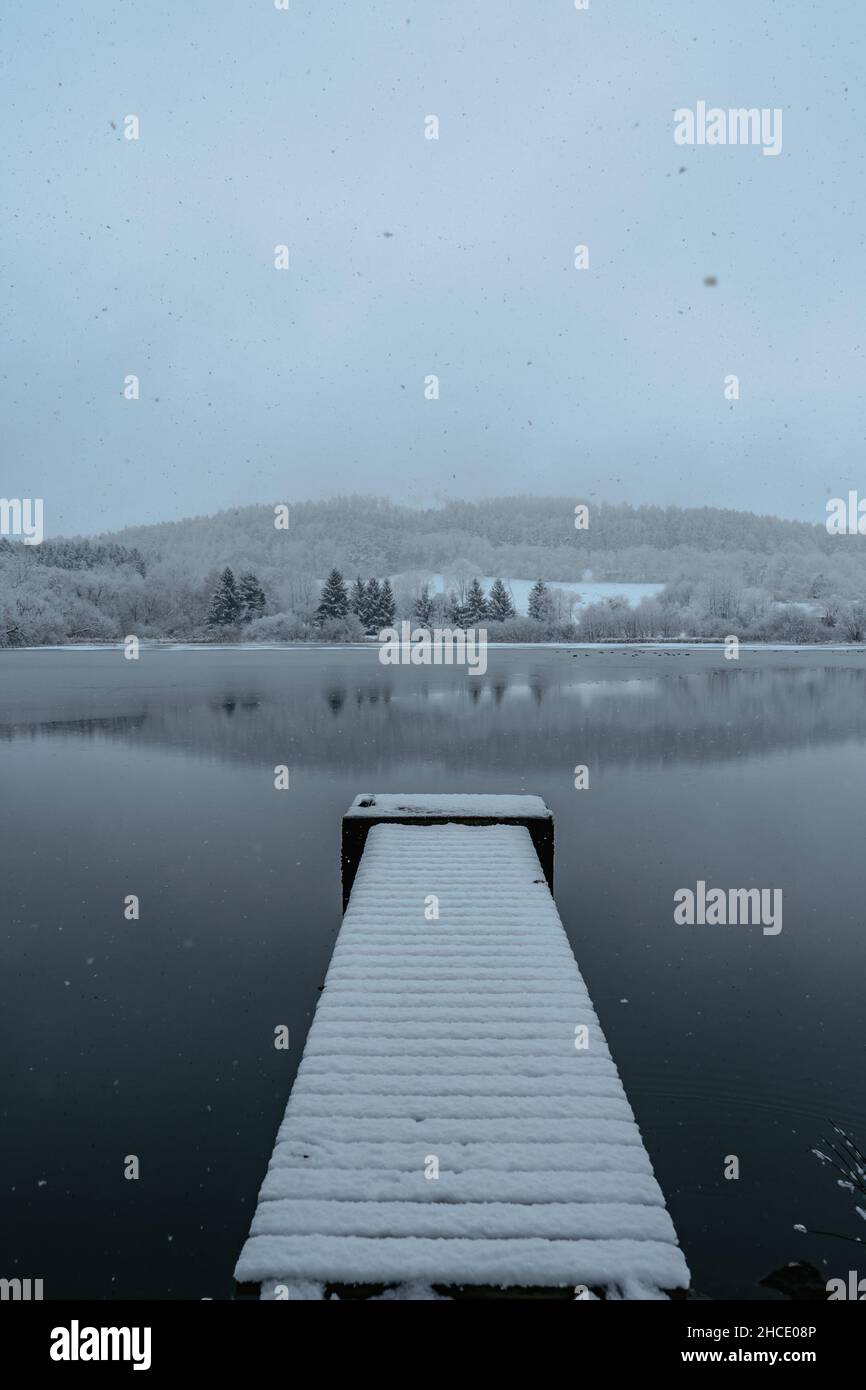 Molo di legno sul lago coperto di neve fresca. Stagno d'inverno con piccolo molo al Morny Morning.Foggy paesaggio nuvoloso riflesso in acqua. Inverno bianco Foto Stock