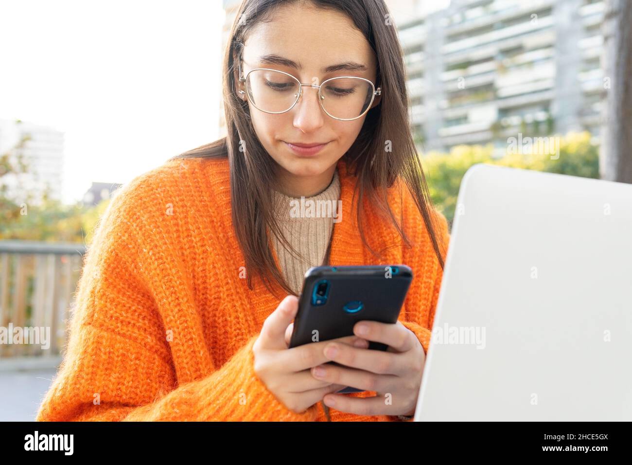 Freelance femminile focalizzata che legge i messaggi sul telefono cellulare mentre lavora con il laptop in remoto nel parco Foto Stock