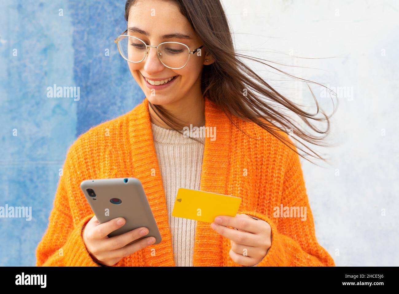 Da sotto di ragazza allegra in occhiali e cardigan arancione lavorato a maglia a pagamento per l'acquisto online con carta di plastica su smartphone Foto Stock