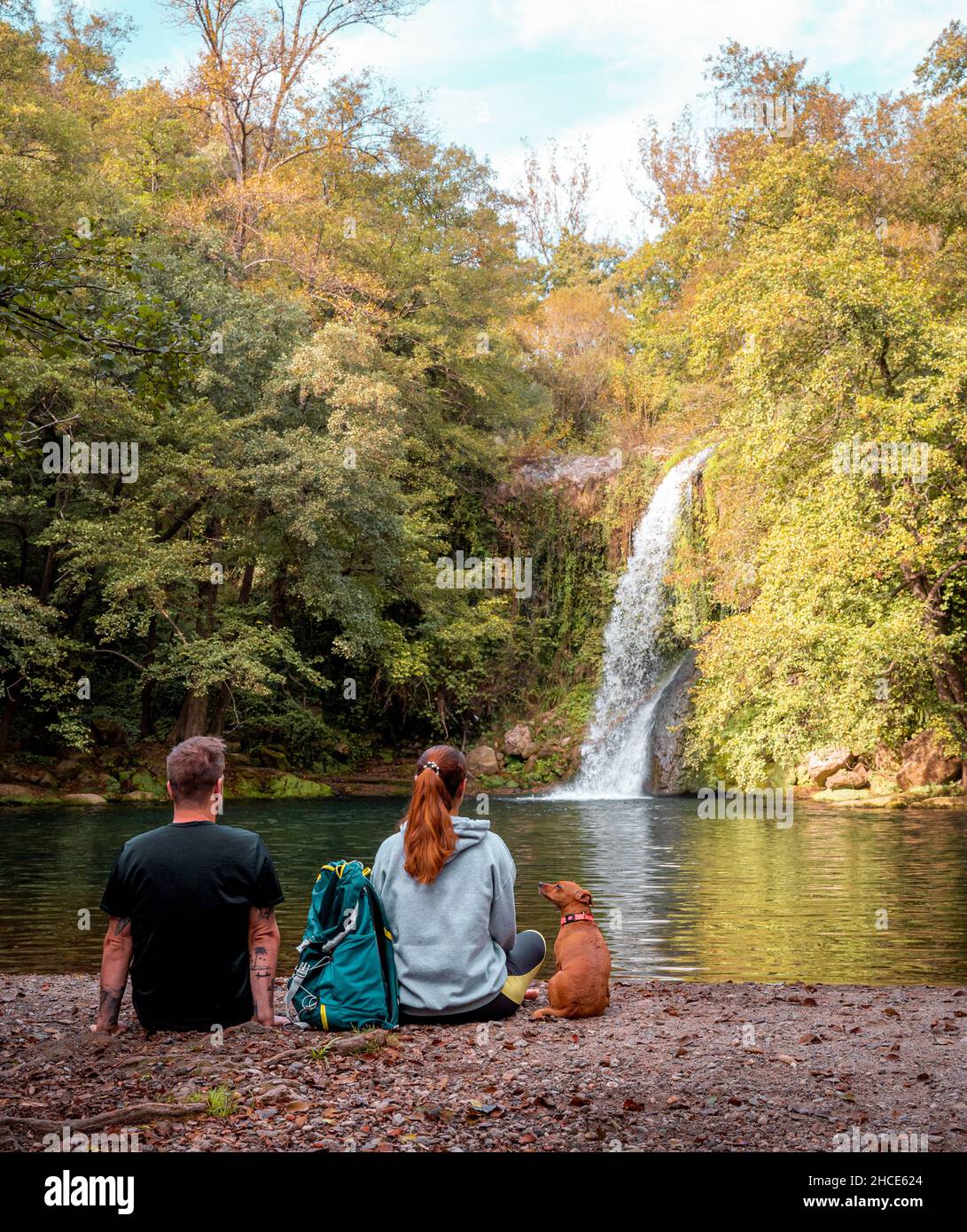 Vista posteriore di coppia anonima seduta con dogon riva del fiume e godere di vista della cascata in Gorg de Santa Margarida Foto Stock