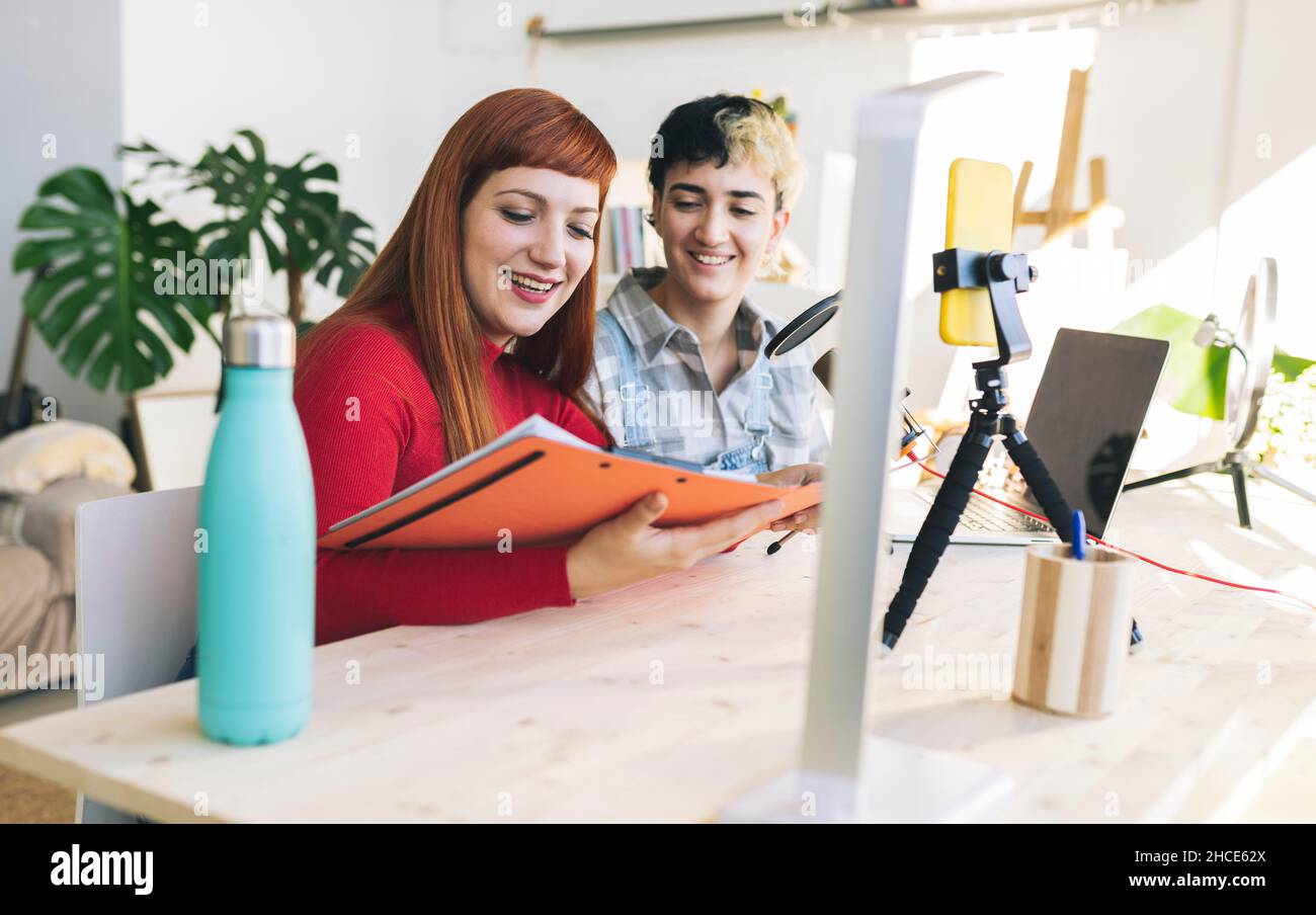 Allegre amici femminili seduti a tavola con documenti e microfono durante la ripresa di podcast su smartphone nella stanza luminosa di casa Foto Stock