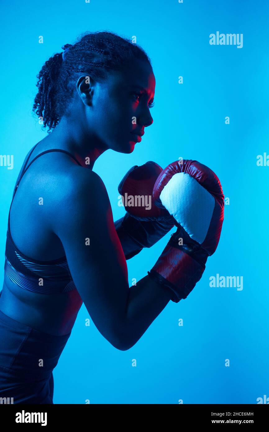 Vista laterale del combattente nero femminile in guanti da boxe in piedi in luce neon blu e tecnica di punzonatura di allenamento Foto Stock