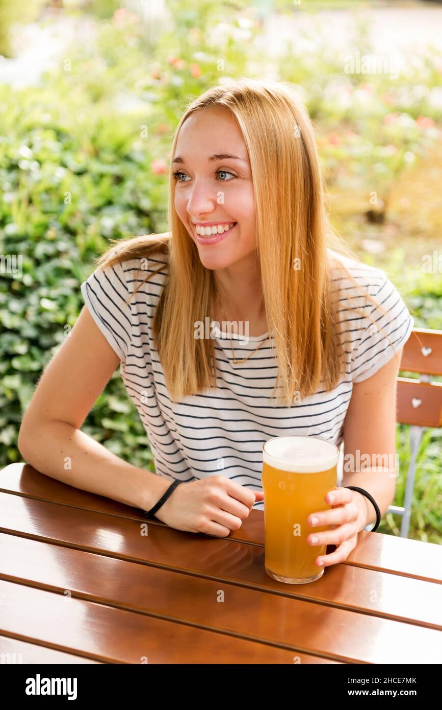 Positiva giovane donna dai capelli biondi in vestito casual seduta a tavola di legno con vetro pieno di fredda schiuma di birra lager e sorridente mentre godendo l'estate Foto Stock