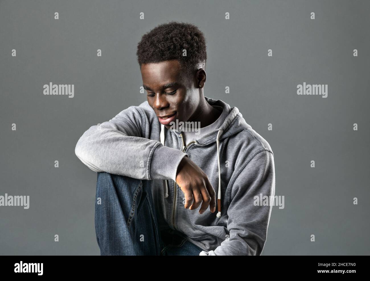 Positivo giovane nero modello maschio con capelli neri Afro scuro in felpa con cappuccio alla moda e jeans sorridenti e guardando giù mentre si siede su sfondo grigio in studi Foto Stock