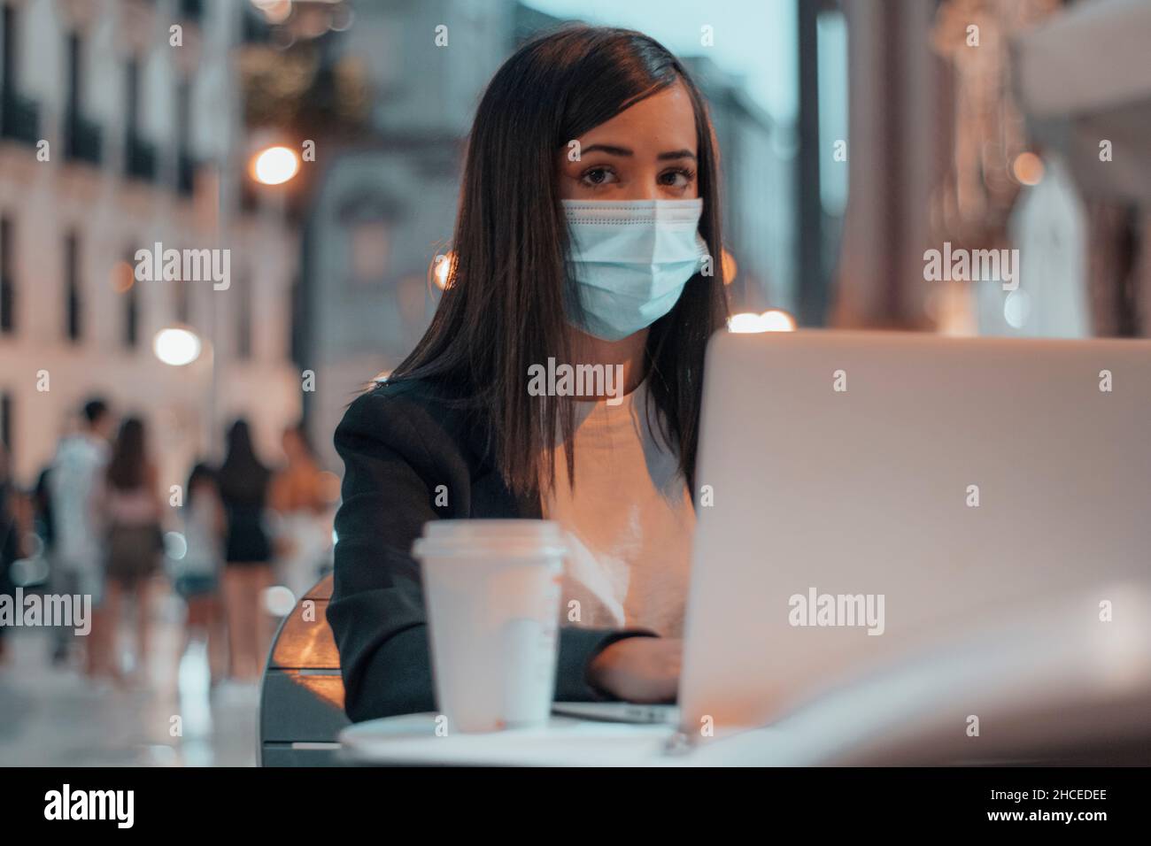 Segretaria di lavoro duro di capelli scuri con la maschera seduta nella piazza della città vicino ad un caffè con il suo laptop Foto Stock