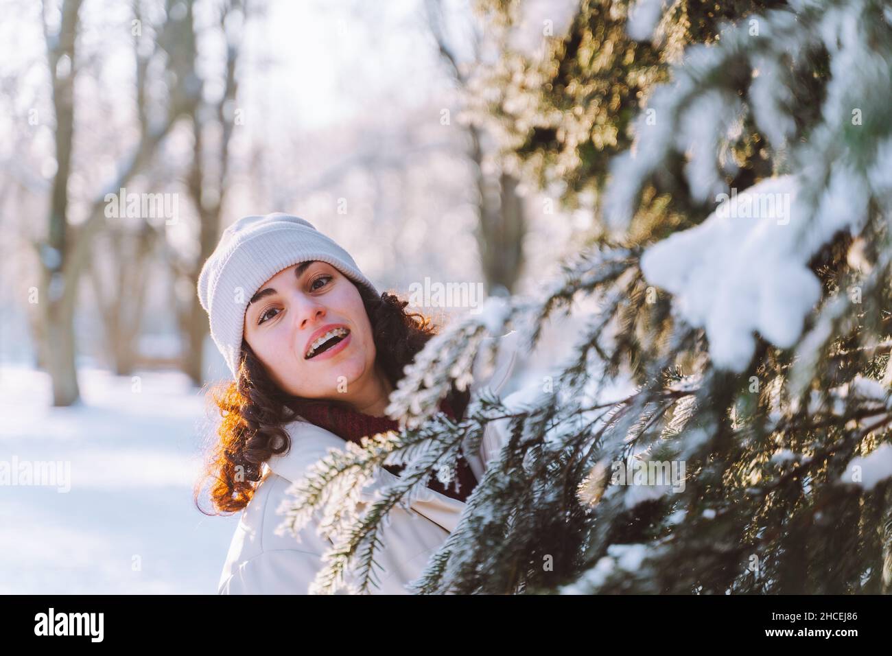 Giovane donna sorridente curly cammina nel parco invernale e nascondersi dietro l'albero di Natale coperto di neve. Emozioni positive, freschezza, vacanze. Foto Stock