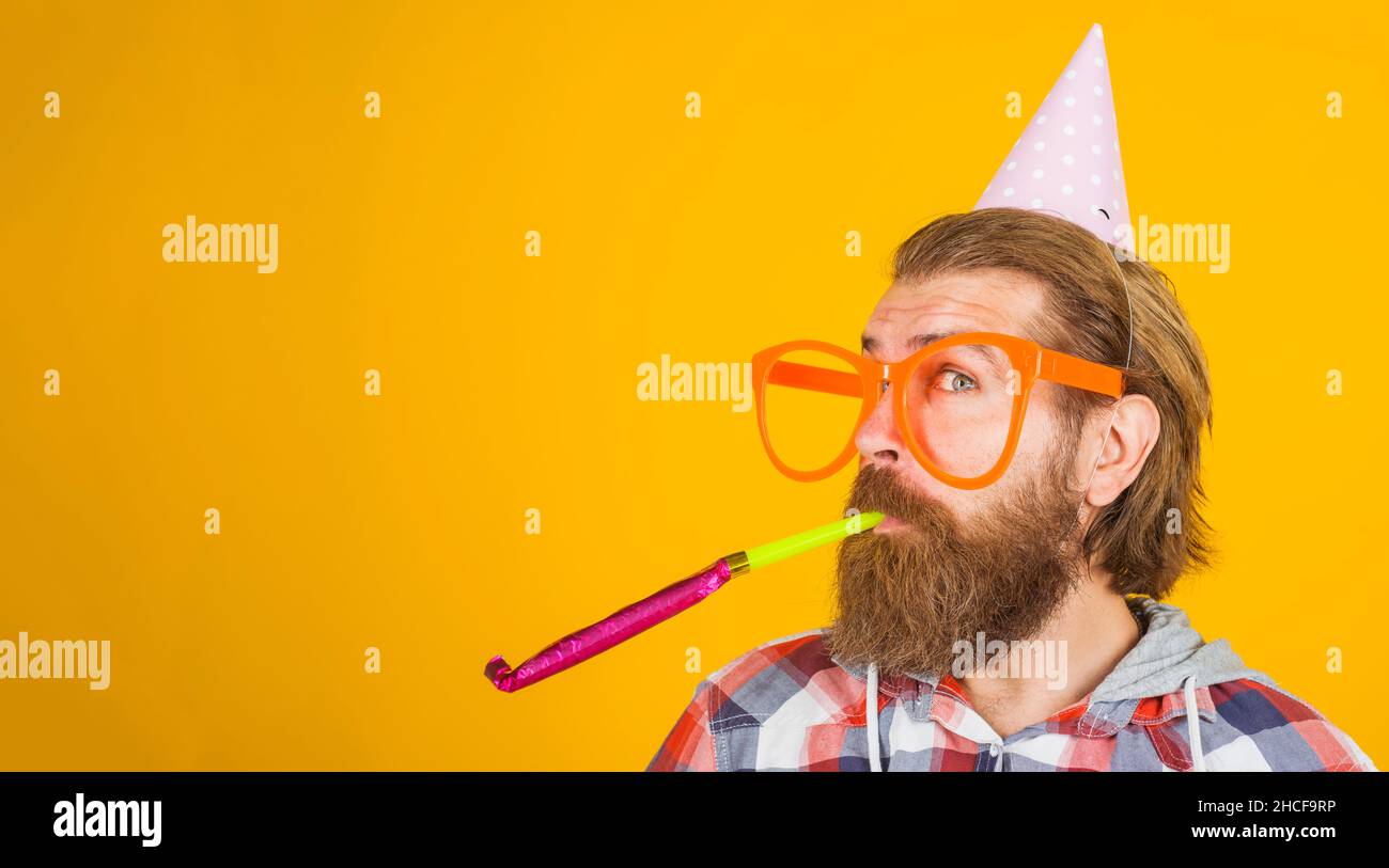Uomo bearded nel cappello di partito soffia noisemaker. Ragazzo felice in bicchieri grandi in berretto cono di compleanno. Spazio di copia. Foto Stock