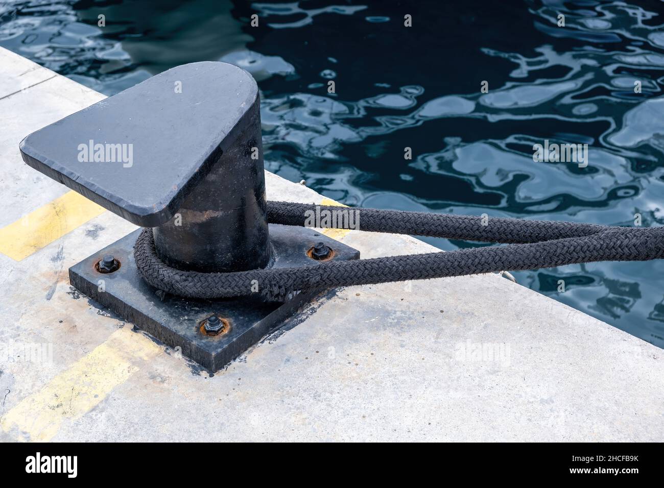Corda di ormeggio della nave intorno ad un bullard del porto sul molo del porto, sfondo ondulato dell'acqua di mare, spazio di copia. Palo in metallo nero ancorato al pavimento in cemento della banchina Foto Stock