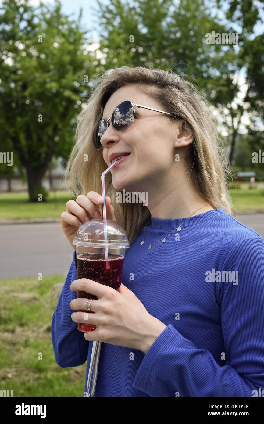 Bella ragazza bionda in occhiali da sole beve un cocktail effervescente attraverso una paglia e sorrisi Foto Stock