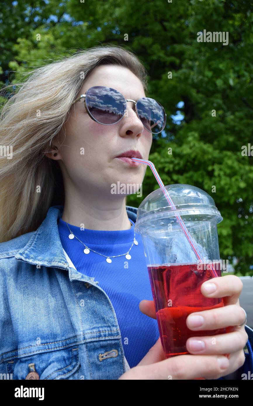 Ragazza bionda attraente in occhiali da sole beve un cocktail effervescente attraverso una paglia Foto Stock