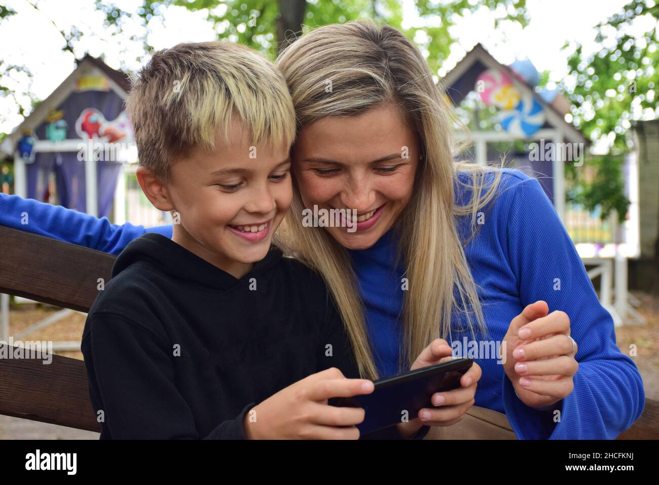 Attraente ragazzo bianco e la sua bella mamma bionda guardare il telefono cellulare e sorridere felicemente Foto Stock