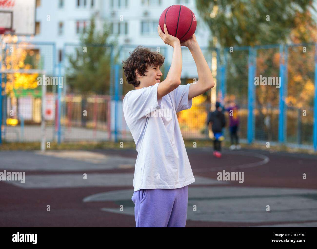 Adolescente carino in felpa con cappuccio viola che gioca a basket. Ragazzo giovane con palla imparare dribble e sparare sul campo della città. Hobby per bambini, Foto Stock