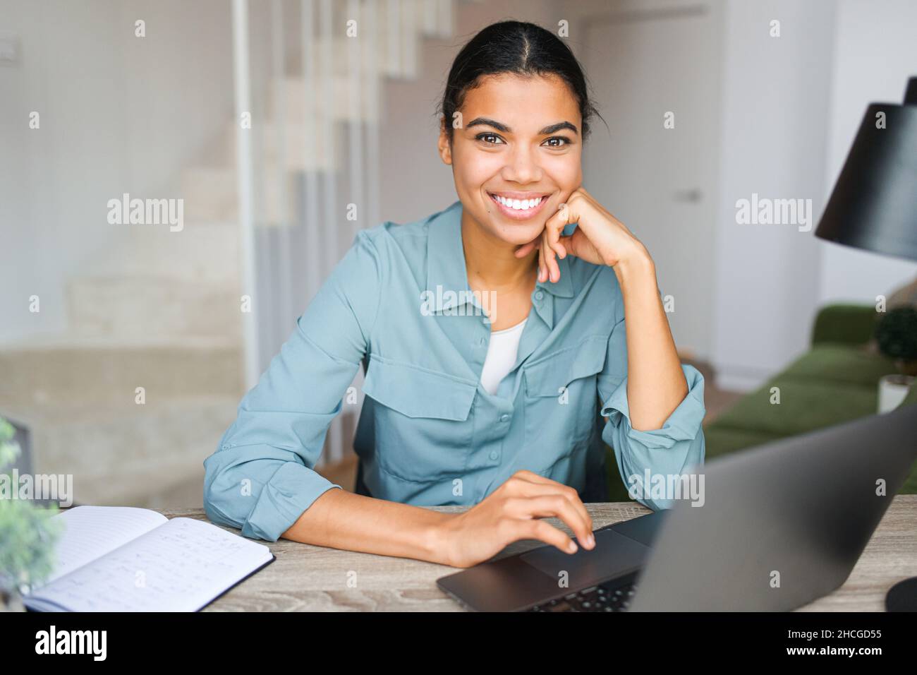Sorridente giovane donna che lavora da casa. Si prepara a parlare alla videochiamata mentre si siede al tavolo davanti al laptop. Attraente ragazza afro-americana Foto Stock