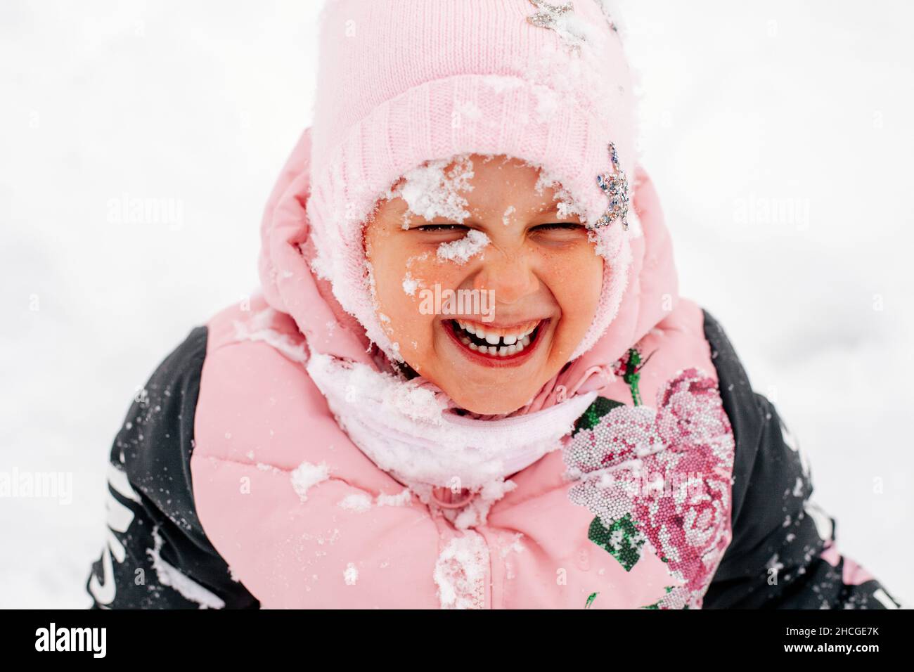 Primo piano foto di femmina russo ragazzo sorridente con denti con il viso coperto di neve con gli occhi chiusi indossando abiti rosa inverno nel parco. Sorprendente Foto Stock