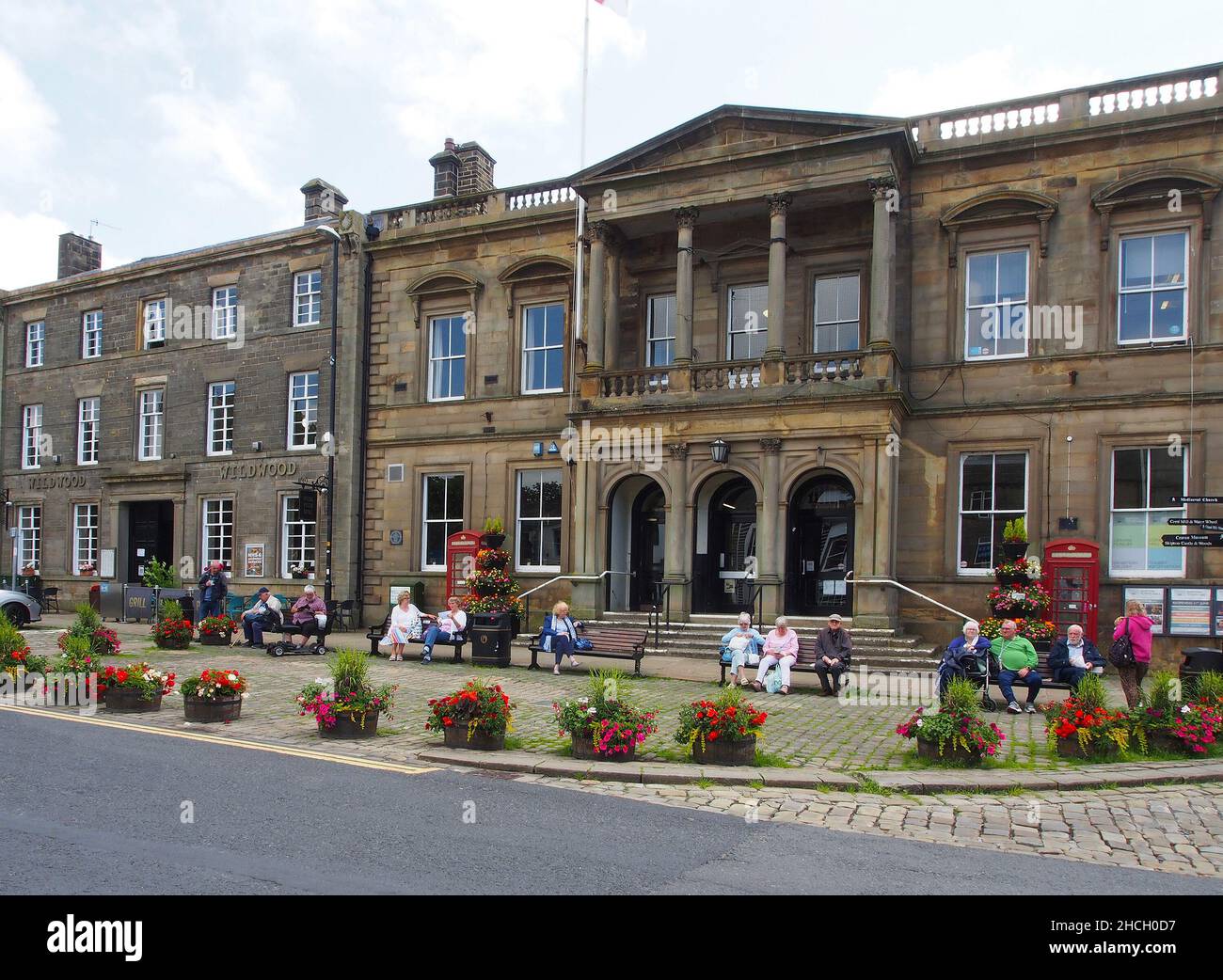 Gli anziani godono del sole seduto su panchine fuori dal municipio di Skipton, North Yorkshire, Inghilterra, con fiori in vasche in primo piano. Foto Stock