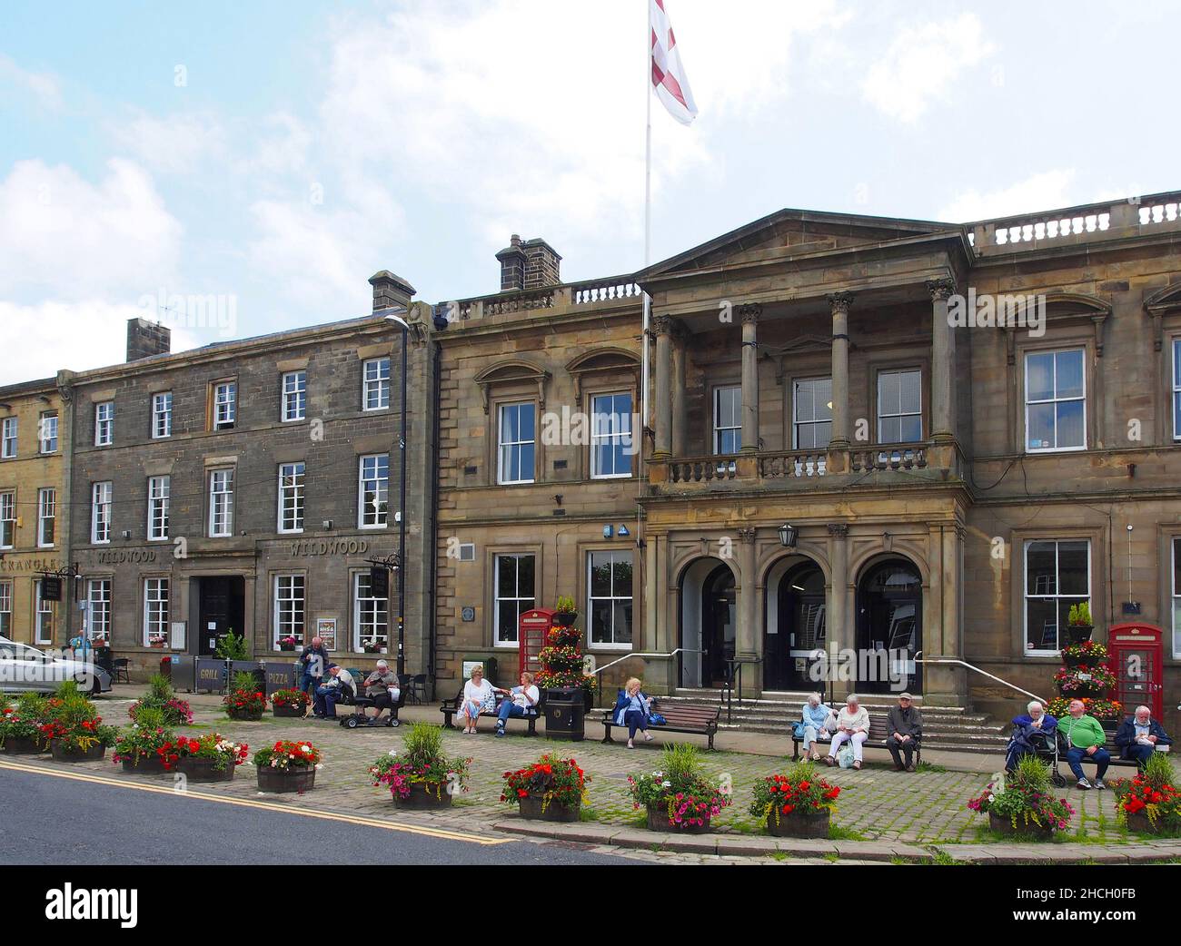 Gli anziani godono del sole seduto su panchine fuori dal municipio di Skipton, North Yorkshire, Inghilterra, con fiori in vasche in primo piano. Foto Stock