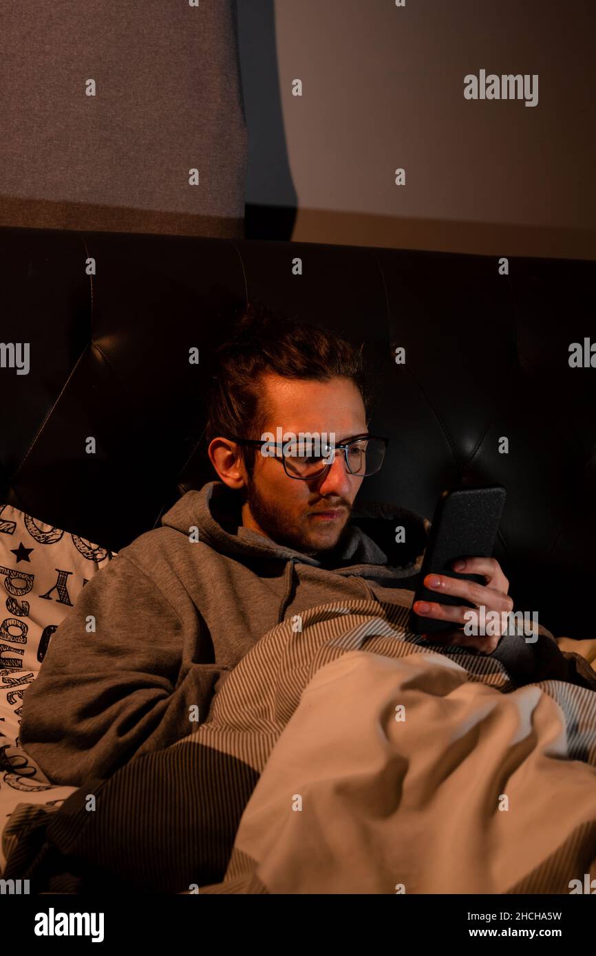 Un giovane addicted ai mezzi sociali che controlla il suo telefono prima di andare a letto, l'aggiunta, i mezzi sociali, dopo il concetto oscuro Foto Stock
