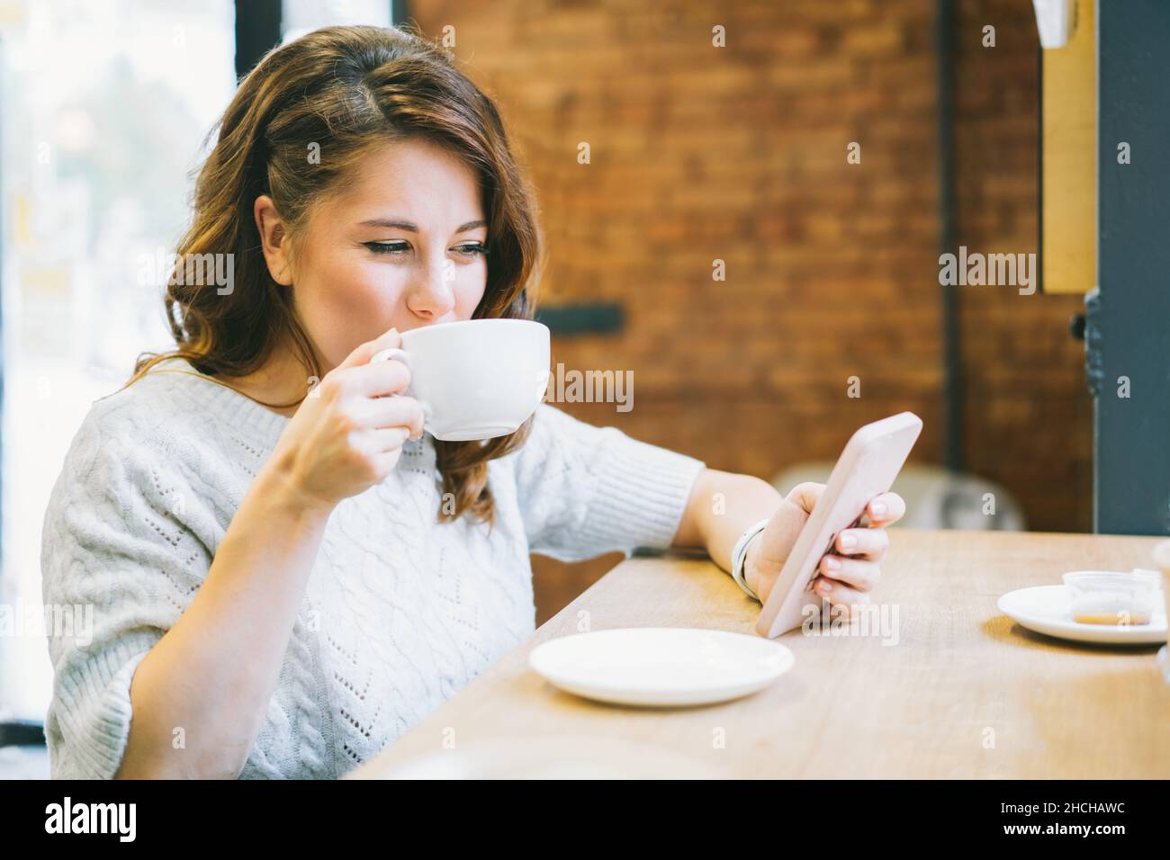 Giovane donna con tazza di caffè in mano messaggi, lettura, SMS, guardare video, scorrimento di social network, prendere appunti, fare acquisti online al telefono Foto Stock