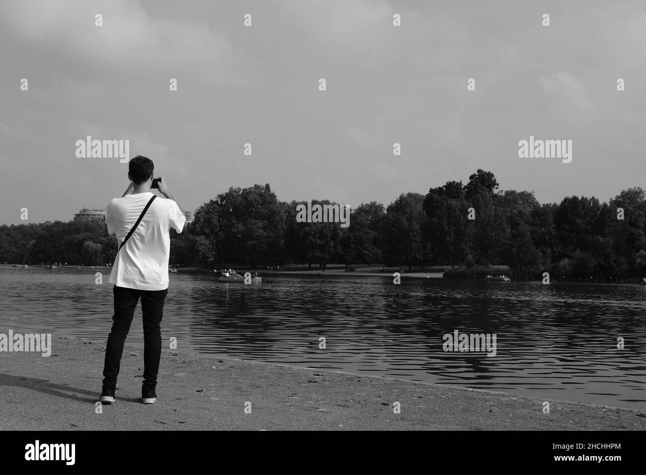 Giovane uomo con ritorno alla macchina fotografica fotografando barche da diporto su Hyde Park Serpentine London UK. In bianco e nero Foto Stock