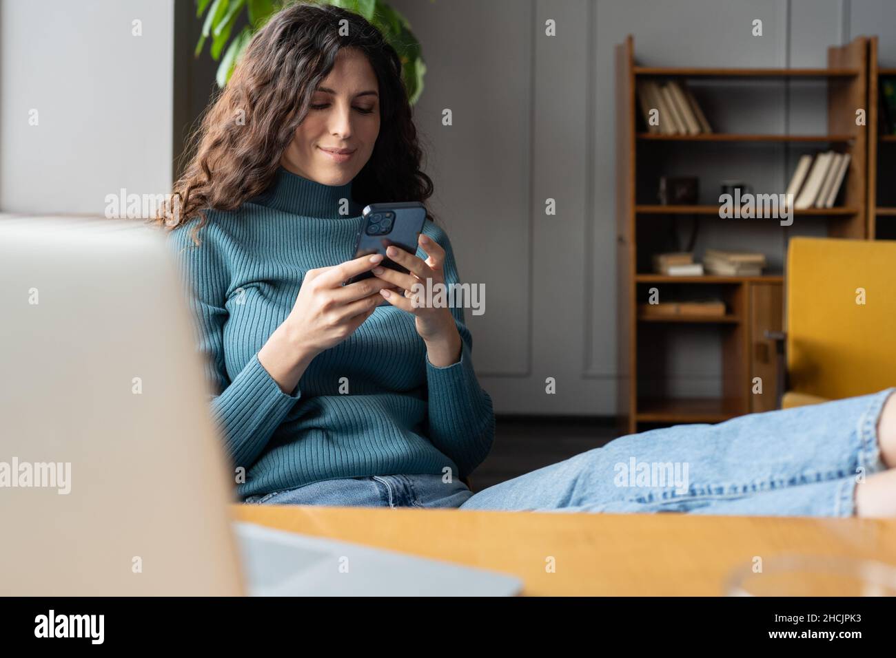 Giovane donna sorridente che tiene lo smartphone chiacchierando online mentre si rilassa in un accogliente ufficio domestico Foto Stock