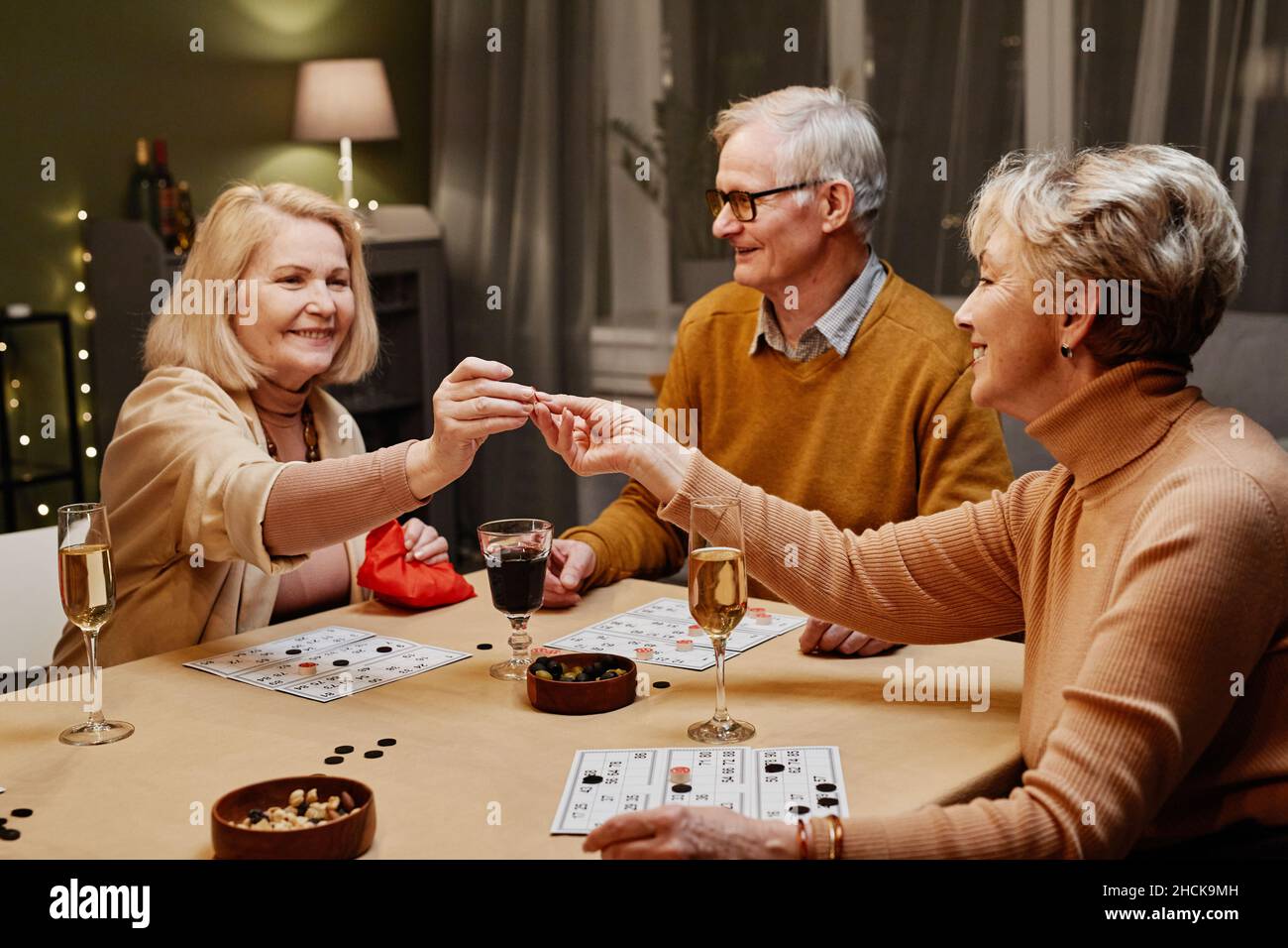 Due donne caucasiche anziane e uomo seduto al tavolo di legno con snack e bevande durante il party e il gioco da tavolo Foto Stock