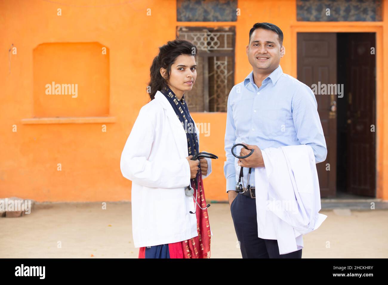 Ritratto di giovane uomo indiano sorridente e femmina Dottore tenuta stetoscopio in piedi guardando la macchina fotografica con emozioni positive contro backgroun arancione Foto Stock